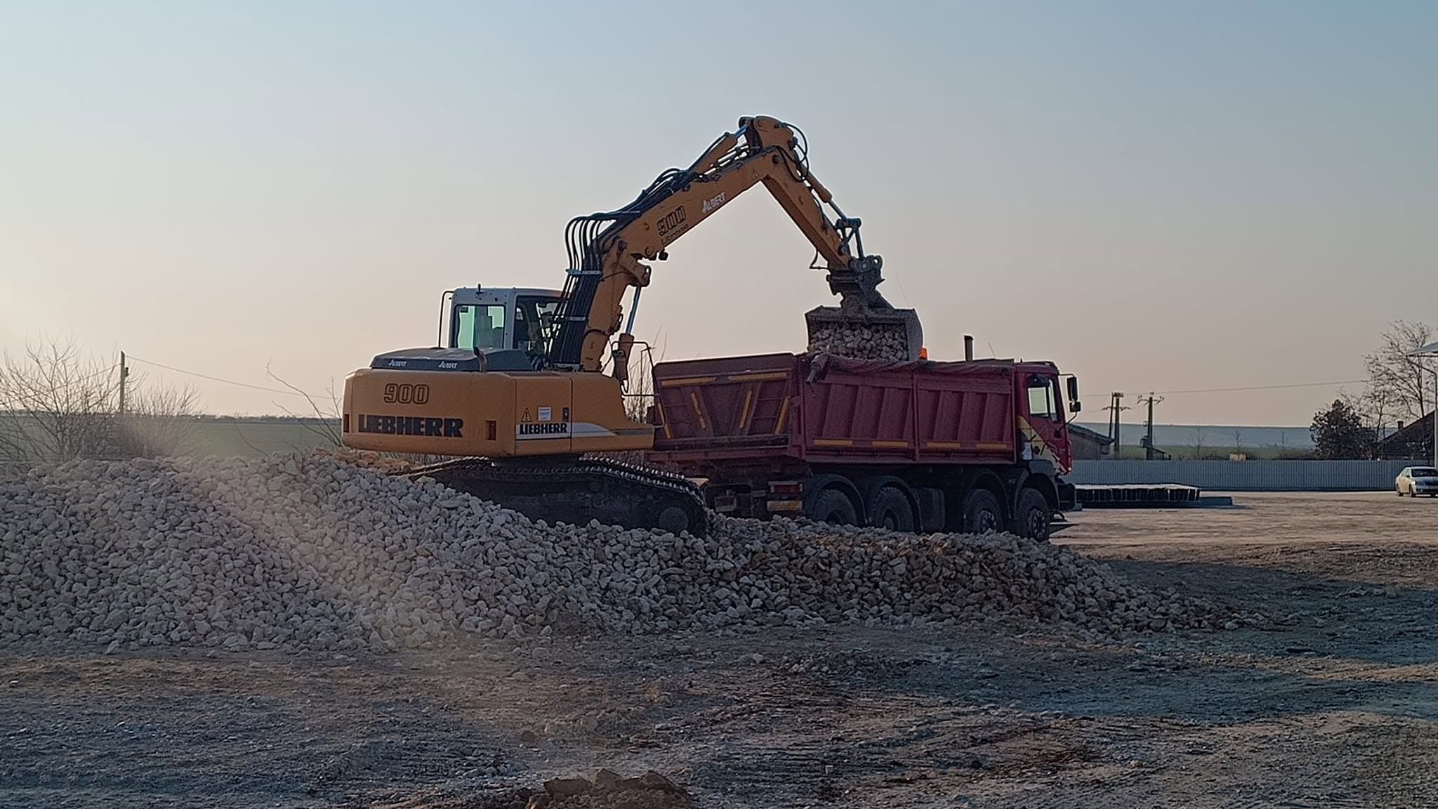 Excavator încărcând o autobasculantă pe un șantier, spre seară