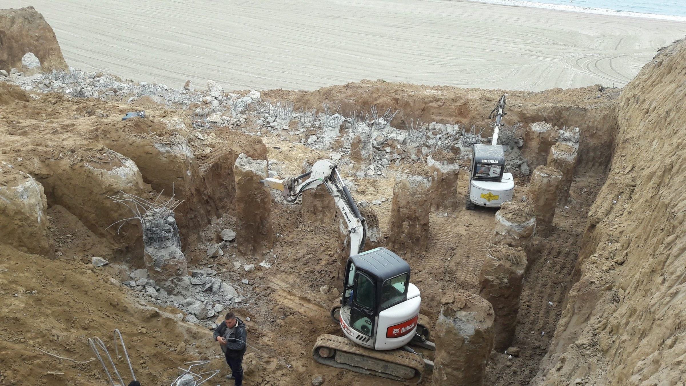 Piloți forați cu armătură, două miniexcavatoare în groapa de fundație, plaja și marea în fundal